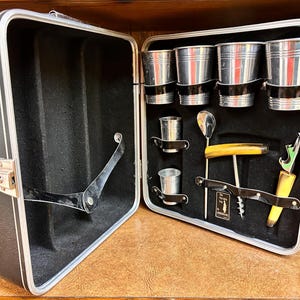 May include: A vintage black and silver bar kit in an open suitcase. The interior has a black lining and holds several small metal cups, a spoon, a corkscrew, and a bottle opener. The case is on a wooden surface.