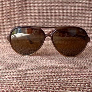 May include: Brown aviator sunglasses with a double bridge and tinted lenses. The sunglasses have a classic teardrop shape and a brown frame. The lenses reflect the surrounding environment.