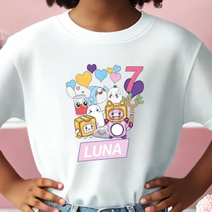 May include: A white t-shirt with a colorful cartoon design featuring a group of characters and the number 7. The shirt has a pink rectangle with the name "LUNA" printed on it.