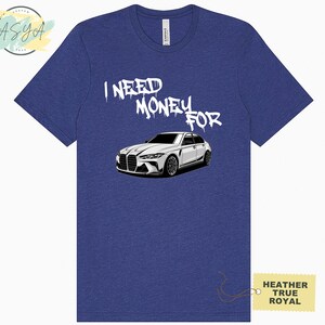 May include: A blue heather t-shirt with the text "I NEED MONEY FOR" in white spray paint lettering. A white car is pictured below the text. The text "HEATHER TRUE ROYAL" is on a yellow tag in the bottom right corner.