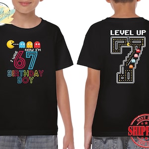 May include: Black t-shirt with a colorful Pac-Man design and the text "I WAS 6 NOW I'M 7 BIRTHDAY BOY" on the front. The back features a pixelated "7" with "LEVEL UP" above it. A fun birthday shirt.