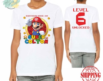 May include: White t-shirt with a cartoon character graphic and the text "SUPER COLTON". The front of the shirt also has the text "BIRTHDAY MODE: ON". The back of the shirt has the text "LEVEL 6 UNLOCKED".