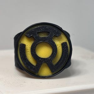 May include: A circular ring featuring a black frame and a yellow center with a raised black symbol resembling a stylized lantern logo. The object has a textured surface finish consistent with 3D printing, showing a clear contrast between the black and yellow components.