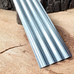 May include: A silver corrugated metal sheet with a slightly curved edge. The sheet is lying on a wooden surface.