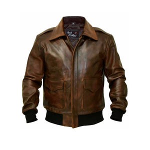 Puede incluir: Una chaqueta bomber de cuero marrón con cuello, puños y cintura de canalé negros. La chaqueta tiene cierre de cremallera y dos bolsillos de solapa en la parte delantera. La etiqueta de la chaqueta dice "Real Leather".