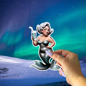 May include: A sticker of a mermaid with white hair and a green tail holding a blue drink with a straw. The mermaid is wearing a green seashell bra. The sticker is on a hand against a background of a starry night sky with green aurora borealis.