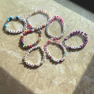 May include: A collection of beaded bracelets in various colors. Some bracelets feature words like "GIRLIE", "MARATHON", and "RUNNERS" spelled out with letter beads. Other bracelets have colorful beads and numbers. The bracelets are arranged on a light-colored surface.