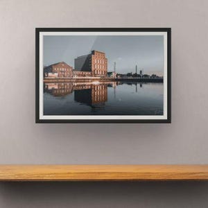 May include: A framed landscape photograph featuring a multi-story brick industrial building situated along a calm body of water. The structure is reflected in the water, with a clear sky above and a thin white mat inside a black frame.