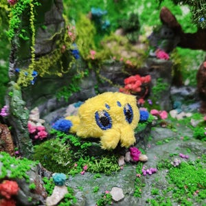 May include: A plush yellow creature with blue accents and large, embroidered eyes rests on a mossy surface. The background features a miniature forest scene with rocks, greenery, and colorful flowers, creating a whimsical setting.
