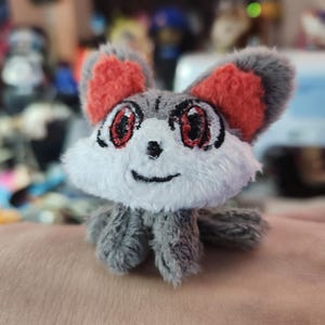 May include: A handmade plush toy of a small, cartoonish creature. It has gray fur, red-tipped ears, and large eyes with red and black details. The creature's face is white, and it has a small, smiling mouth. The toy is designed to resemble a mouse or similar animal.