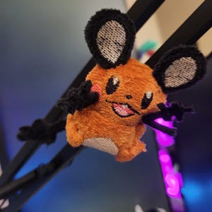 Può includere: Un peluche di un personaggio di topo dei cartoni animati con pelliccia arancione, orecchie nere e braccia nere. Il topo ha guance rosa e una bocca rosa cucita. Lo sfondo è sfocato, con accenni di blu e viola.