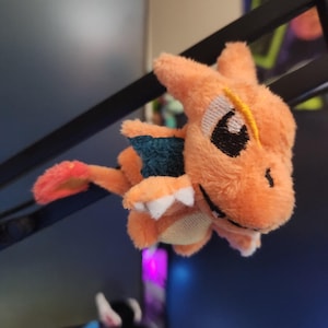 Può includere: Un peluche arancione di una creatura simile a un drago con pancia bianca, occhi neri e una striscia gialla. La creatura ha un'ala blu e una coda rossa. Il giocattolo è appeso a un oggetto nero.