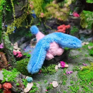 May include: A plush toy bird with blue wings and a pink body is set in a miniature forest scene. The scene includes green moss, small pink and red flowers, and a backdrop of rocks and foliage.