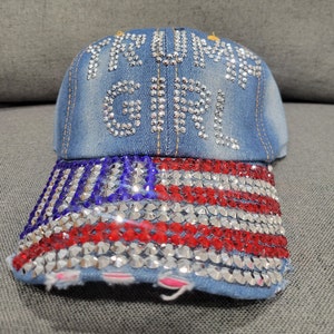 May include: A blue denim baseball cap with a red, white, and blue American flag design on the front. The cap is decorated with rhinestones and says "Trump Girl" in rhinestones.
