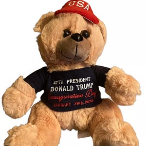 Könnte beinhalten: Ein beigefarbener Teddybär, der ein blaues Shirt mit dem Text "47th President Donald Trump Inauguration Day January 20th, 2021" und eine rote Mütze mit dem Text "USA" in Weiß trägt.