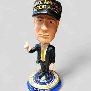 May include: A bobblehead figurine of Donald Trump wearing a black baseball cap with the text "Make America Great Again" in yellow letters. The figurine is wearing a black suit with a yellow tie and is giving a thumbs up. The base of the figurine is blue with a gold presidential seal and the text "Donald Trump" in white letters.