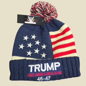 Puede incluir: Un gorro de punto azul marino con un pompón rojo, blanco y azul. El gorro presenta estrellas blancas bordadas, rayas rojas y blancas, y la palabra "TRUMP" con "MAKE AMERICA GREAT AGAIN" y "45-47" bordado en rojo.