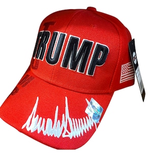 May include: Red baseball cap with the name "TRUMP" in large, black, block letters. The cap features an American flag patch and a signature design on the brim. A black tag is attached to the side.