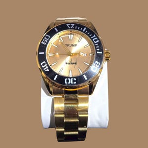May include: Gold-toned wristwatch with a black bezel and a gold dial. The watch features the name "TRUMP" on the dial, along with a signature. The watch has a gold metal band and is displayed on a white surface.