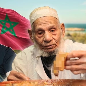 Op de afbeelding: Een man in een witte gewaden en een witte hoed eet een maaltijd met zijn handen. Hij zit aan een tafel met een man in een zwart shirt en een man in een bruin shirt. De man in het zwarte shirt kijkt met een verraste uitdrukking naar de camera. De man in het bruine shirt eet met zijn handen. De achtergrond is een wazig beeld van een strand en een blauwe lucht.