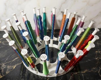 May include: A white, circular organizer filled with colorful crochet hooks. The hooks have various colored handles, including green, blue, red, and orange. Each hook has a white top with a number on it, such as "01" or "21".