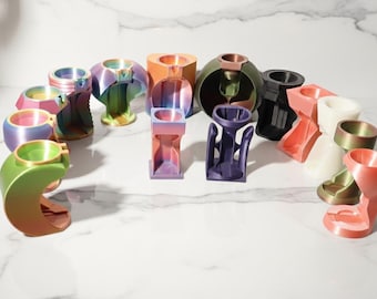 May include: A collection of colorful, 3D-printed vases in various shapes and sizes. The vases feature a gradient of colors, including green, pink, purple, and orange. Some have a metallic finish, and all are displayed on a white marble surface.