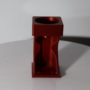 May include: A red, geometric candle holder with a square top and a rectangular base. The design features curved cutouts on the sides, creating a modern and artistic look. The candle holder is made of a solid material and has a metallic finish.