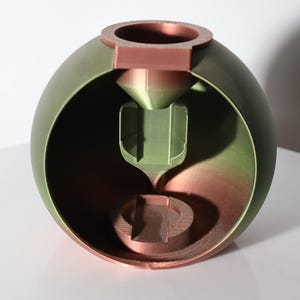 May include: A 3D-printed, spherical object with a cutaway revealing an interior structure. The exterior is a gradient of green and brown, with a copper-colored ring at the top. The interior features a funnel-like design.