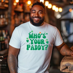 Pode incluir: Uma camiseta branca com um gráfico verde que diz "Who's Your Paddy?" com desenhos de trevos ao redor do texto. A pessoa que está usando a camiseta está sorrindo e segurando um copo de cerveja.