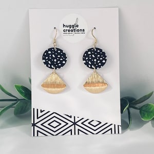 May include: Dangle earrings featuring a black circular disc with a white speckled pattern attached to a gold-toned fan-shaped metal piece with a brushed texture. The earrings are displayed on a white card with the text huggle creations.