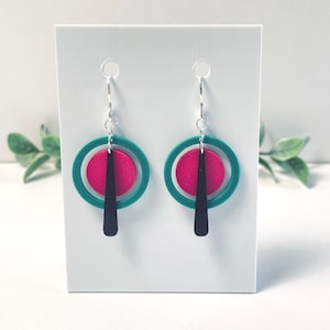 May include: A pair of drop earrings featuring a silver-toned hook, a teal circular ring, a smaller hot pink circular disc, and a black elongated teardrop-shaped pendant. The earrings are displayed on a white rectangular card for presentation.