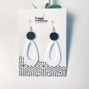 May include: Dangle earrings featuring a small black textured circle attached to a larger white teardrop-shaped cutout. The earrings hang from gold-toned hooks on a white display card with the text huggle creations and a geometric pattern.