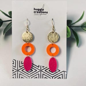 May include: Dangle earrings featuring a vertical arrangement of three shapes: a textured gold-tone circle, a bright orange ring, and a vibrant pink oval. The earrings are attached to gold-tone hooks and displayed on a white card with the text huggle creations.