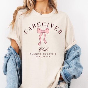 May include: A beige short-sleeved t-shirt featuring a graphic design with a pink bow and the text CAREGIVER CLUB RUNNING ON LOVE & RESILIENCE in dark brown lettering. The shirt is worn with a blue denim jacket and denim shorts.
