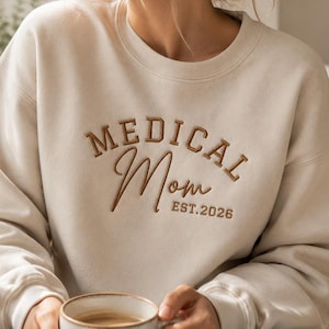May include: A beige crewneck sweatshirt featuring brown embroidered text that reads MEDICAL Mom EST.2026. The garment has long sleeves and a ribbed neckline, cuffs, and hem, providing a simple and clean look for casual daily wear.