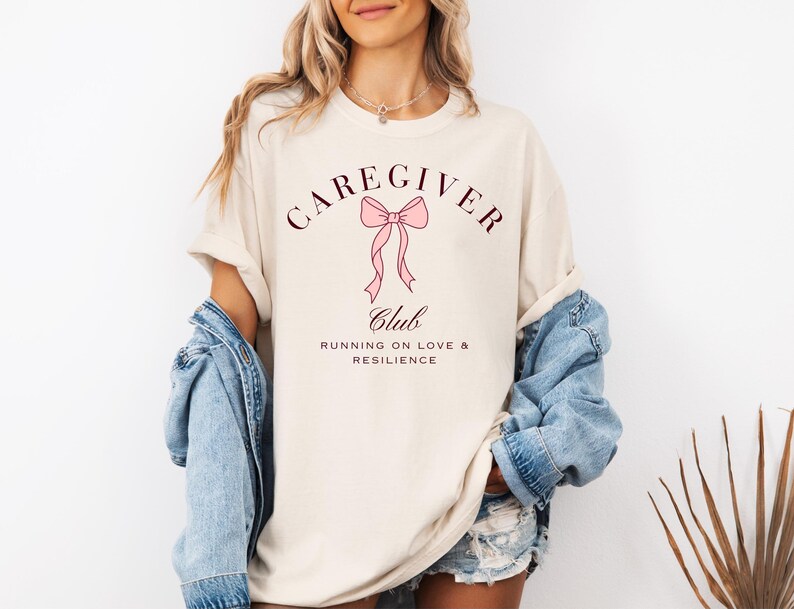 May include: A beige short-sleeved t-shirt featuring a graphic design with a pink bow and the text CAREGIVER CLUB RUNNING ON LOVE & RESILIENCE in dark brown lettering. The shirt is worn with a blue denim jacket and denim shorts.