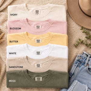 May include: A stack of six short-sleeved crew neck t-shirts in ivory, blossom, butter, white, sandstone, and moss colors. Each shirt features a Comfort Colors brand label on the inside collar. The shirts are arranged vertically in a neat pile.
