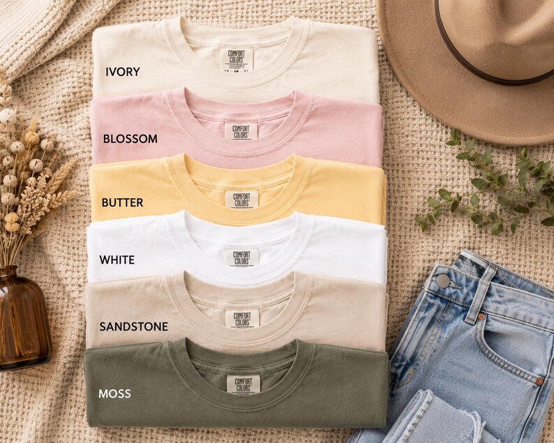 May include: A stack of six short-sleeved crew neck t-shirts in ivory, blossom, butter, white, sandstone, and moss colors. Each shirt features a Comfort Colors brand label on the inside collar. The shirts are arranged vertically in a neat pile.