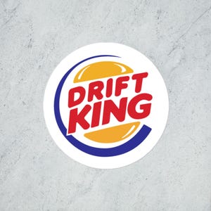 Puede incluir: Una pegatina redonda blanca con el texto "DRIFT KING" en rojo, dentro de un diseño amarillo y azul. El diseño recuerda a un logotipo de comida rápida. La pegatina está sobre un fondo gris.