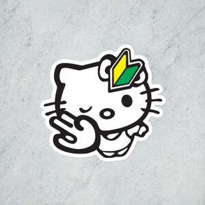 May include: A white sticker featuring a cartoon cat with a black outline, winking and making a hand gesture. The cat has a green and yellow beginner driver symbol on its head. The sticker is on a gray background.