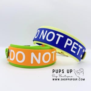 May include: Two dog collars with lime green edges and silver rivets. One collar is orange with white text reading DO NOT, and the other is blue with white text reading DO NOT PET. The text PUPS UP Dog Boutique WWW.SHOPPUPSUP.COM is visible.