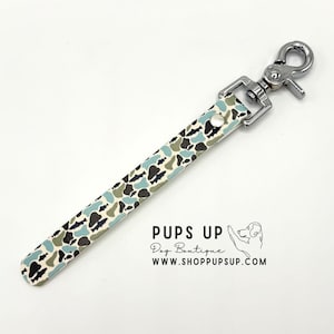 Puede incluir: Accesorio para correa de perro con cierre de plata y correa estampada. La correa presenta un diseño de camuflaje en tonos azules, verdes y negros sobre un fondo crema. El texto "PUPS UP Dog Boutique" y una dirección web son visibles.