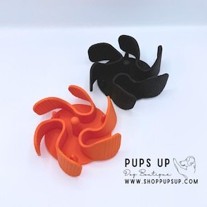 May include: Two dog bowls, one orange and one black, are shown against a white backdrop. The bowls have a distinctive, curved design with a central raised area. The orange bowl is in the foreground, and the black bowl is behind it. The bowls are from Pups Up Dog Boutique.
