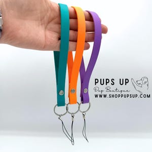 May include: Three colorful dog leash wrist straps in teal, orange, and purple. Each strap has a silver-colored metal ring and a matching silver snap. The straps are held up against a white background. The text "PUPS UP Dog Boutique" is visible.