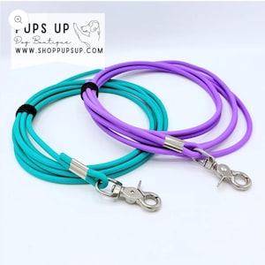 May include: Two dog leashes, one teal and one purple, are displayed. Each leash is made of a thick, rounded cord with a silver-colored clasp and metal accents. The leashes are coiled and connected with a black band.