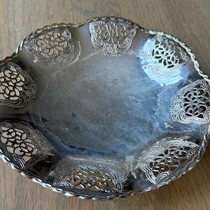 May include: A silver-colored, decorative bowl with a scalloped edge and intricate openwork designs. The bowl has a slightly tarnished appearance, with a reflective surface. The rim features a twisted rope-like detail.