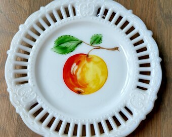 May include: White, decorative plate with a painted apple design. The plate features a detailed, openwork rim and a hand-painted apple with red and yellow hues, green leaves, and a brown stem. A charming piece for display or serving.