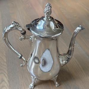 May include: A polished silver-toned teapot featuring an ornate, floral-embossed handle, a matching curved spout, and a decorative finial on the hinged lid. The body of the teapot is smooth and reflective, supported by three detailed, claw-style feet.