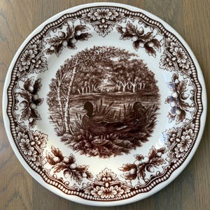 May include: A round ceramic plate featuring a brown and white transferware pattern. The center depicts two ducks near a pond surrounded by trees. The wide rim is decorated with a repeating border of oak leaves and acorns in brown ink.