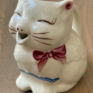 May include: A cream-colored ceramic cat-shaped creamer pitcher featuring a winking expression with painted brown whiskers, a maroon bow tie at the neck, and a thin blue line around the base. The cat has pointed ears and a small open mouth spout.